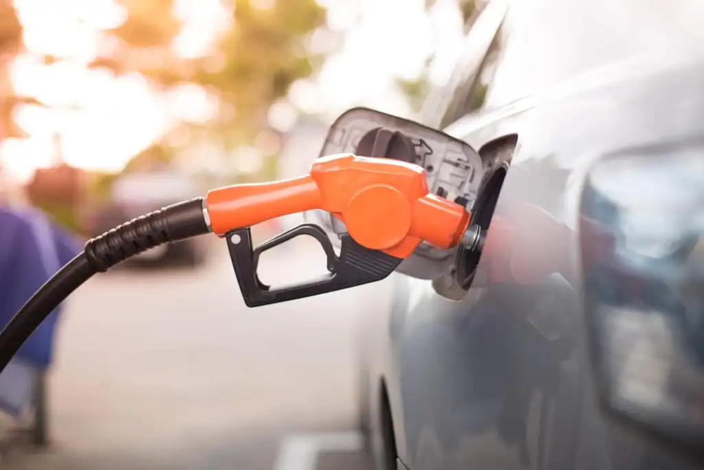 A close-up of an orange fuel pump nozzle inserted into a car’s gas tank, with the car’s fuel cap open and a blurred outdoor background.