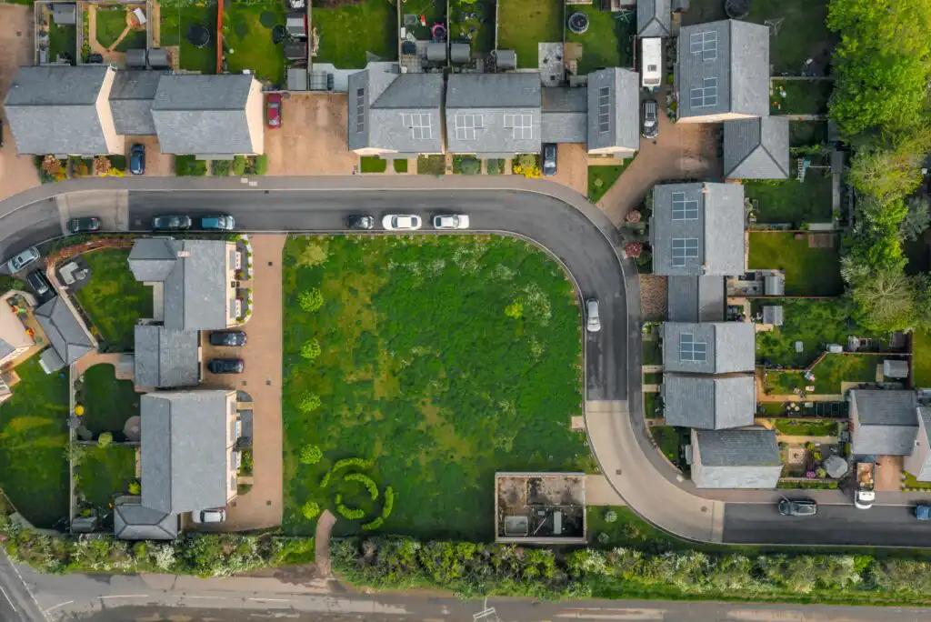 Aerial view of a curved residential street bordered by houses with driveways and gardens, surrounding a central grassy area; several cars are parked along the road and in driveways.
