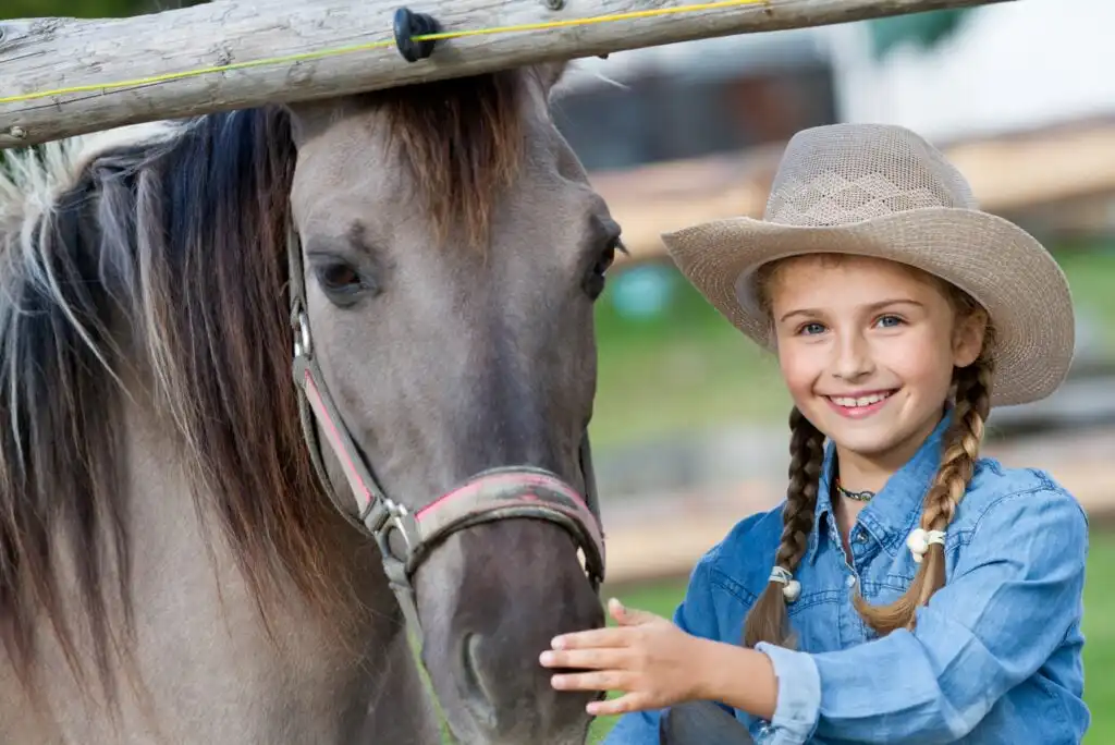 A smiling young girl in a cowboy hat and denim shirt stands next to a brown horse, gently petting its face. The background includes wooden fences and greenery.
