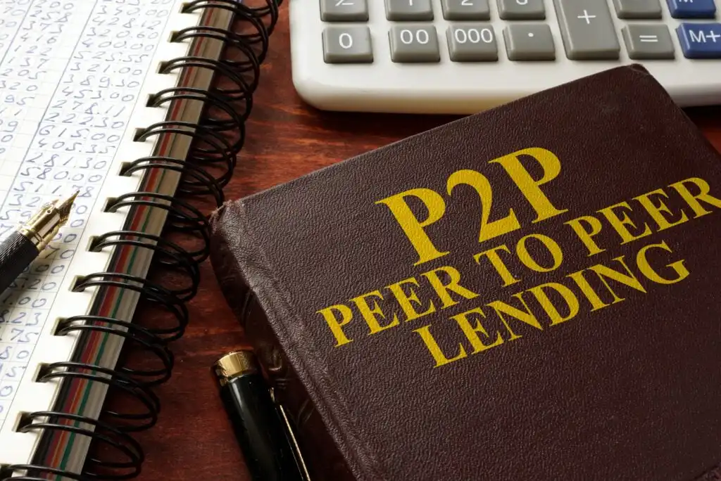 A book titled P2P Peer to Peer Lending lies on a desk beside a notepad with handwritten numbers, a pen, and part of a calculator, suggesting financial or investment planning.