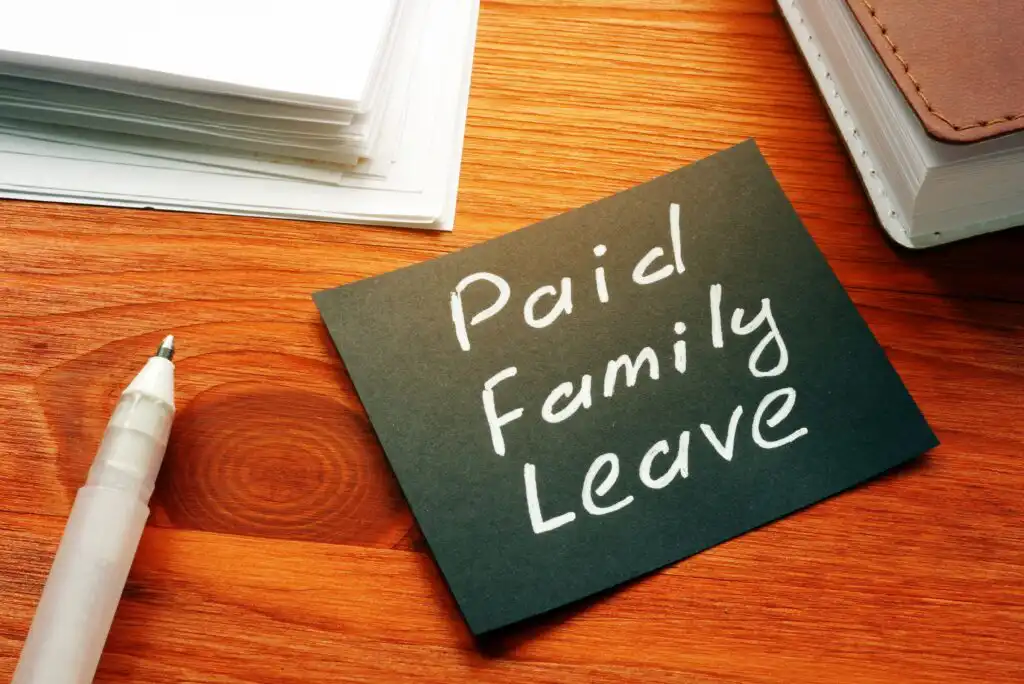 A black card with Paid Family Leave written in white ink, placed on a wooden desk next to a white pen, a stack of papers, and a notebook.
