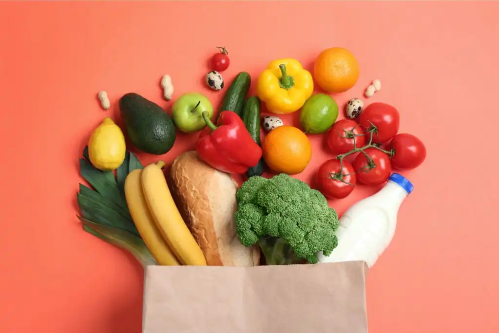 A grocery bag filled with various fresh foods, including bananas, bread, broccoli, avocado, leek, red and yellow bell peppers, tomatoes, cucumber, lemon, oranges, lime, cherry tomatoes, quail eggs, peanuts, and a bottle of milk.