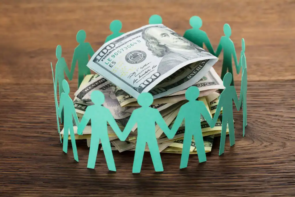 A circle of green paper cut-out people holding hands surrounds a pile of assorted U.S. dollar bills on a wooden surface.