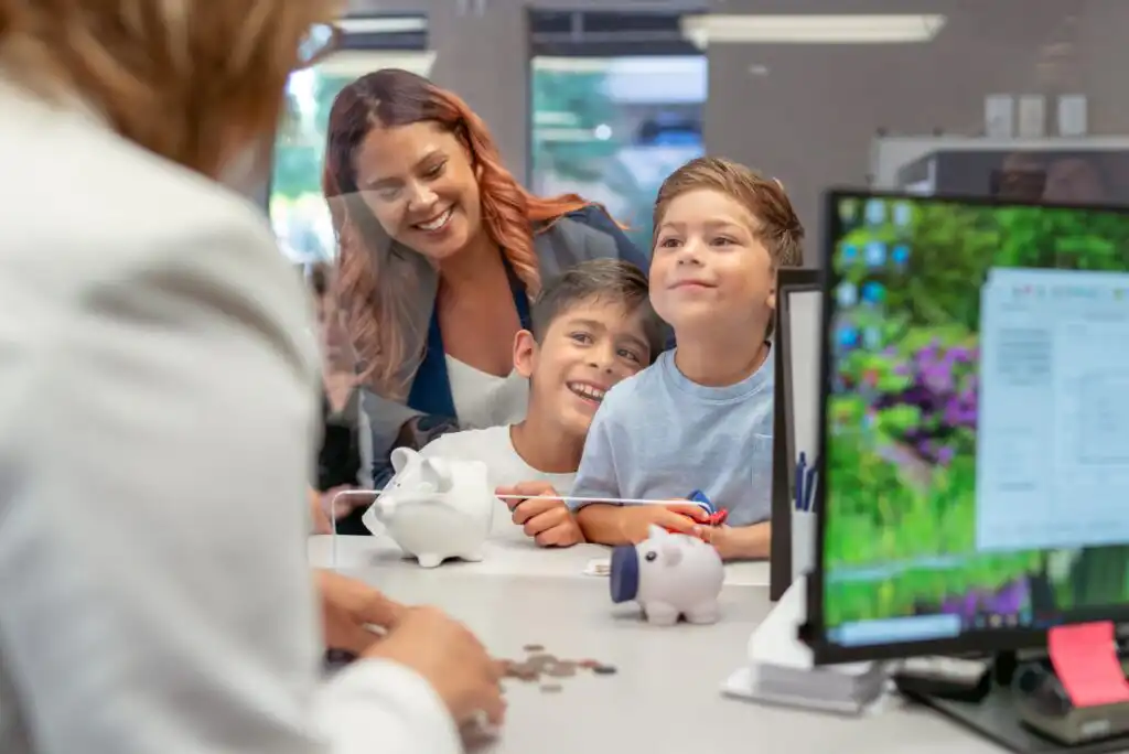 A woman and two young boys smile at a bank teller from across a counter. The boys have toy piggy banks, and coins are visible on the counter. A computer monitor and paperwork are also on the desk.