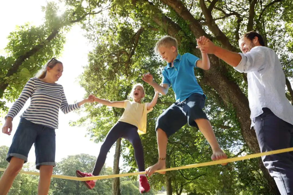 A family of four balances on a slackline outdoors, holding hands for support. Sunlight filters through trees, and the children, one girl and one boy, look excited and happy as parents assist on either side.