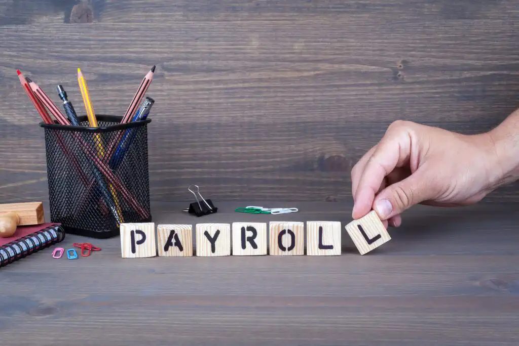 A hand arranges wooden blocks spelling “PAYROLL” on a desk beside a pen holder with pencils and pens, notebooks, and office supplies, all set against a wooden background.