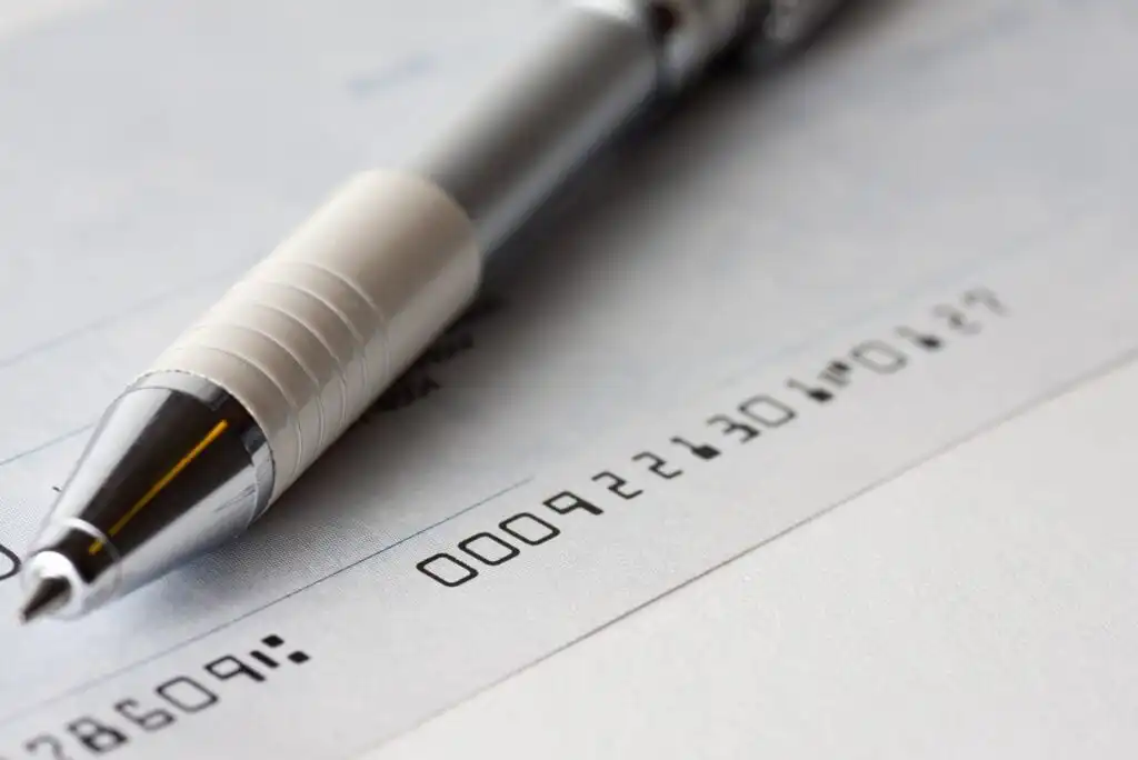 A close-up of a silver and white pen resting on a blank check, with visible numbers and a faint signature line.