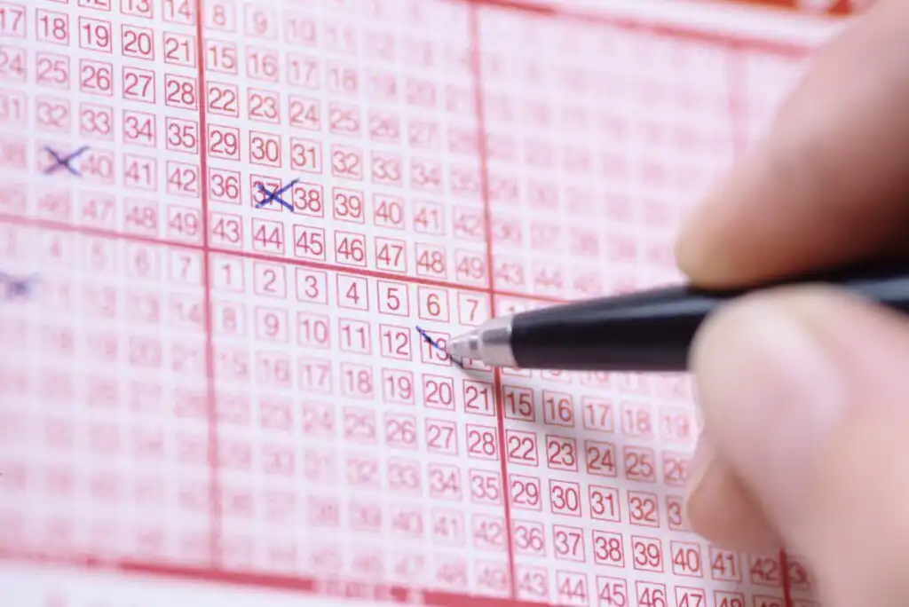 A close-up of a person marking numbers on a lottery ticket with a pen. Several boxes with numbers are visible, some already marked with blue crosses.