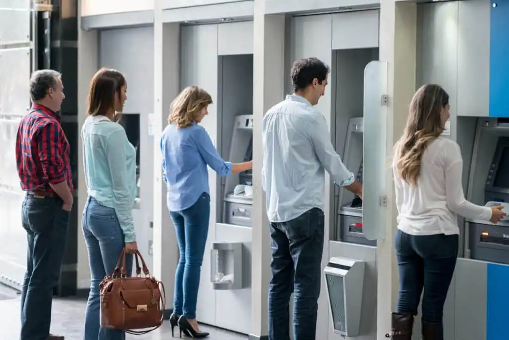 Five people stand in line at ATMs inside a bank, each using a separate machine. The setting is modern, and everyone appears focused on their transactions.