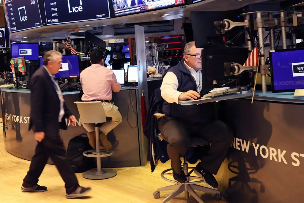 Traders work at their desks with multiple monitors on the floor of the New York Stock Exchange, with one person walking past and financial screens displayed in the background.