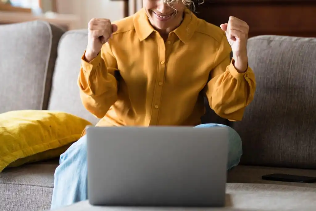 A person sitting on a couch smiles and raises their fists in excitement while looking at a laptop in front of them. They are wearing a mustard yellow shirt and light blue jeans.
