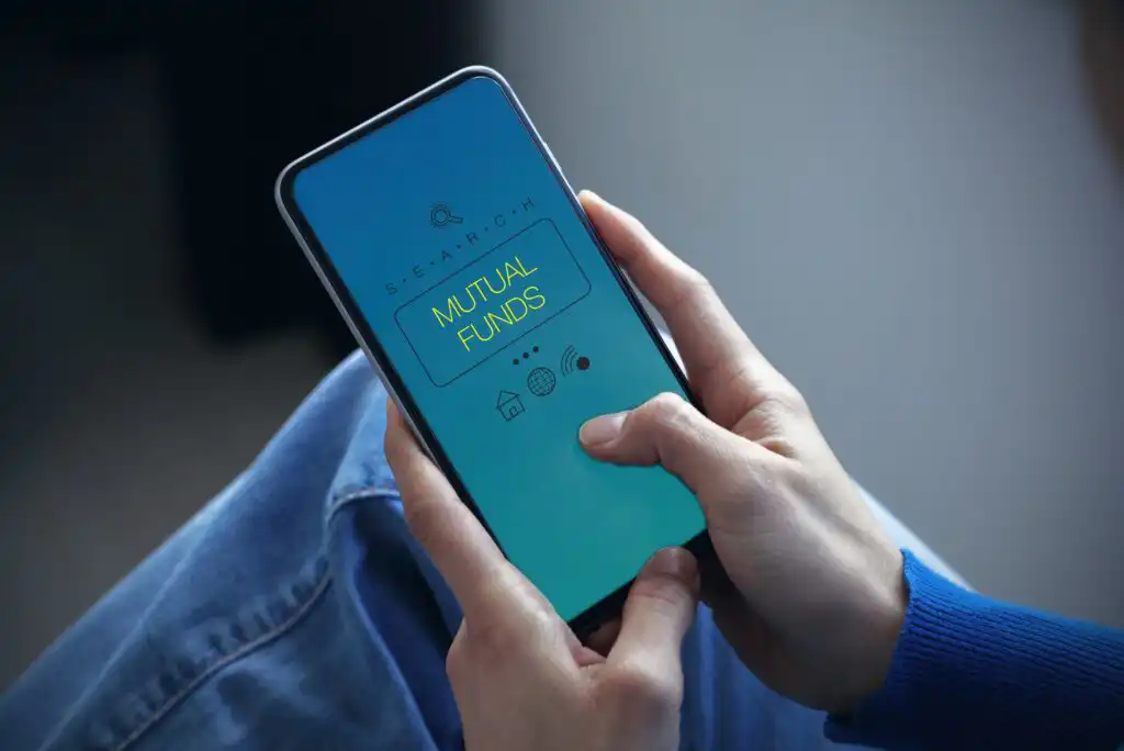A person holds a smartphone displaying a search screen with the words “MUTUAL FUNDS” highlighted, indicating a search or informational app. The person is wearing blue jeans and a blue sweater.