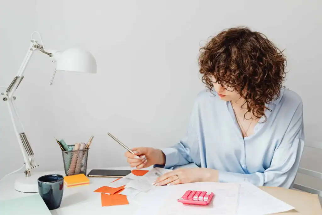 A woman with curly hair in a light blue blouse sits at a desk, reviewing papers and receipts with a pen. A pink calculator, sticky notes, a cup, and a desk lamp are also on the white workspace.