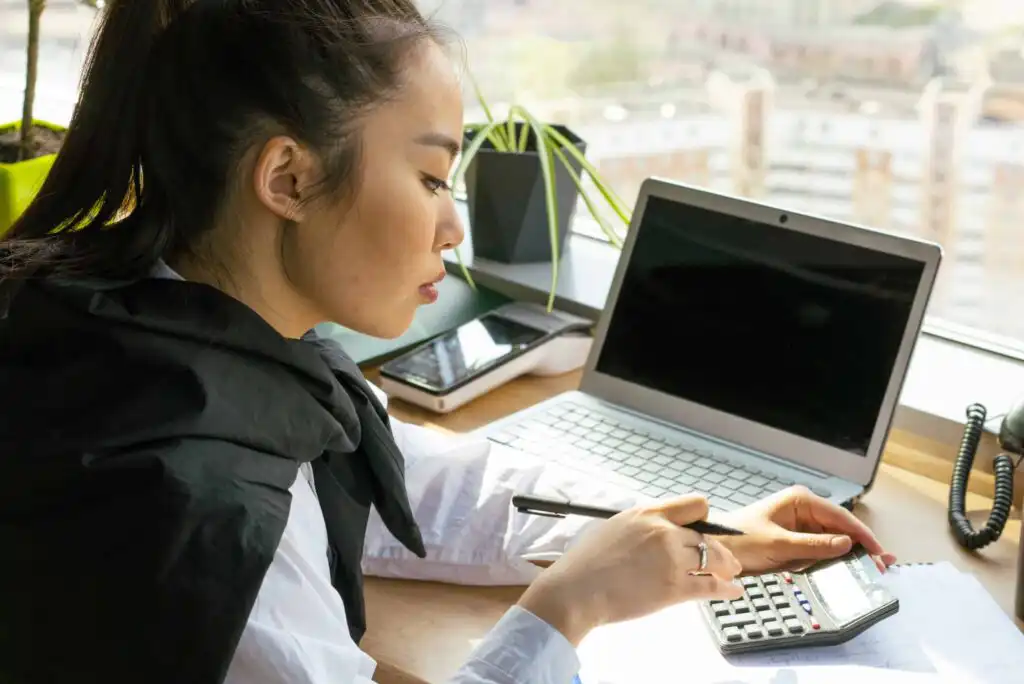A woman sits at a desk by a window, using a calculator and holding a pen, with an open laptop, phone, and potted plant nearby. She appears focused on her work.