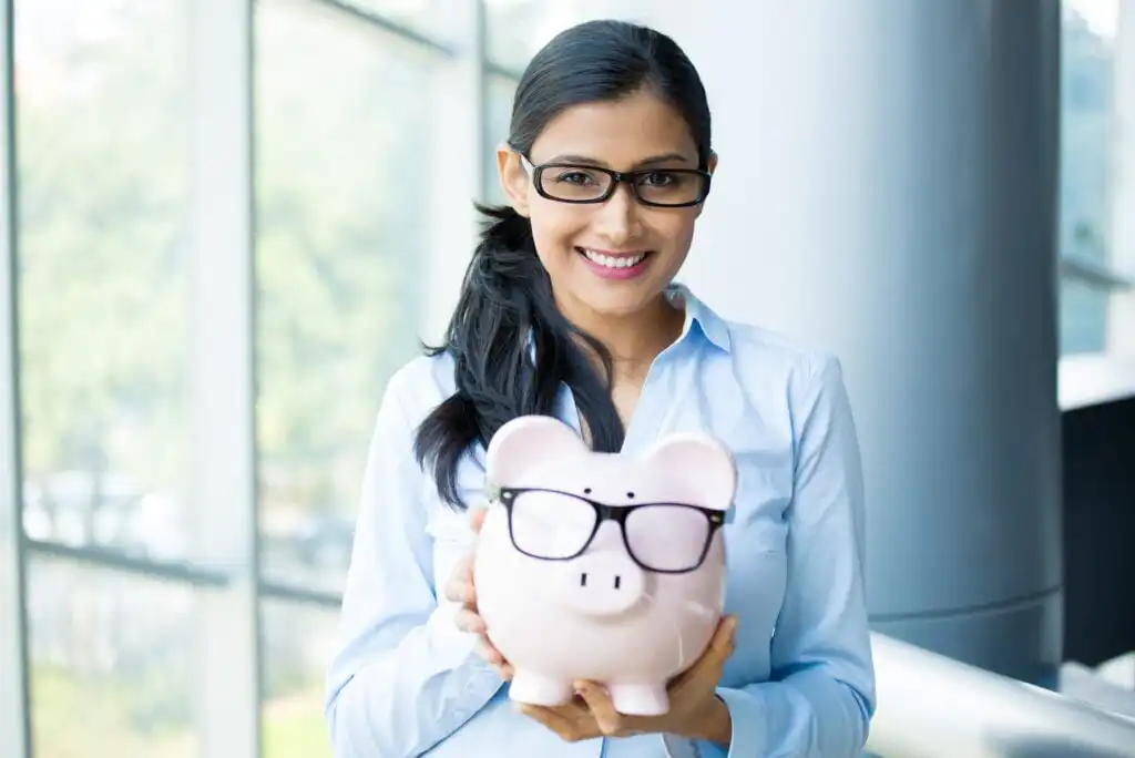 A smiling woman wearing glasses and a light blue shirt holds a pink piggy bank, which is also wearing glasses. She stands in a bright, modern indoor space with large windows in the background.