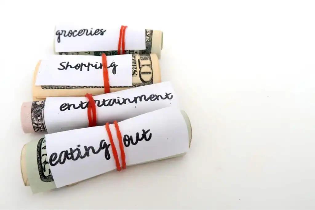 Four rolled-up dollar bills, each labeled with handwritten categories—groceries, shopping, entertainment, and eating out—secured with red rubber bands, representing a budgeting or money allocation system.