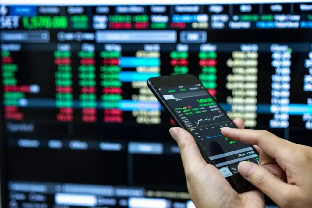 A person uses a smartphone displaying stock market graphs and data, with a large digital stock trading board showing numbers and charts in the background.