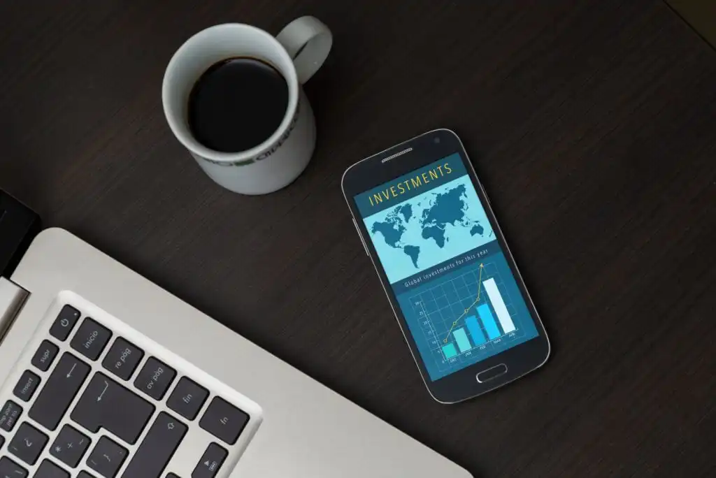 A smartphone displaying an investments chart with a rising bar graph and world map, next to a laptop keyboard and a cup of black coffee on a dark wooden table.