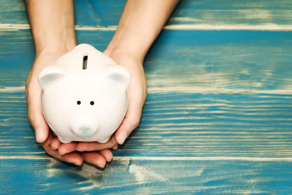 A pair of hands gently holding a white piggy bank on a blue wooden surface.