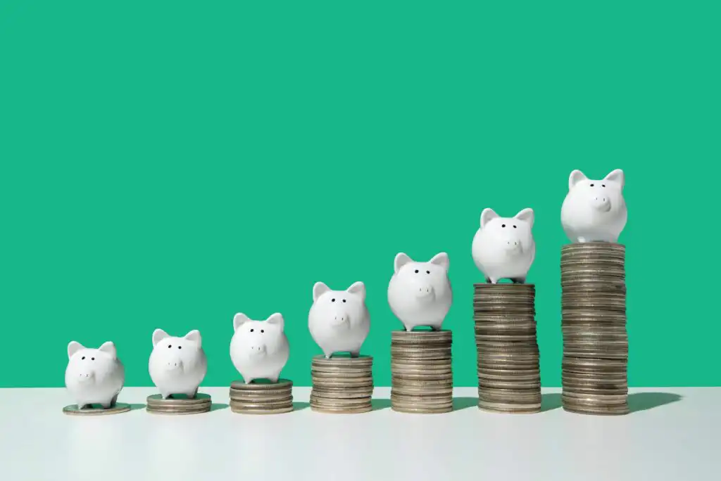Six white piggy banks are lined up on ascending stacks of coins against a green background, symbolizing increasing savings or financial growth.