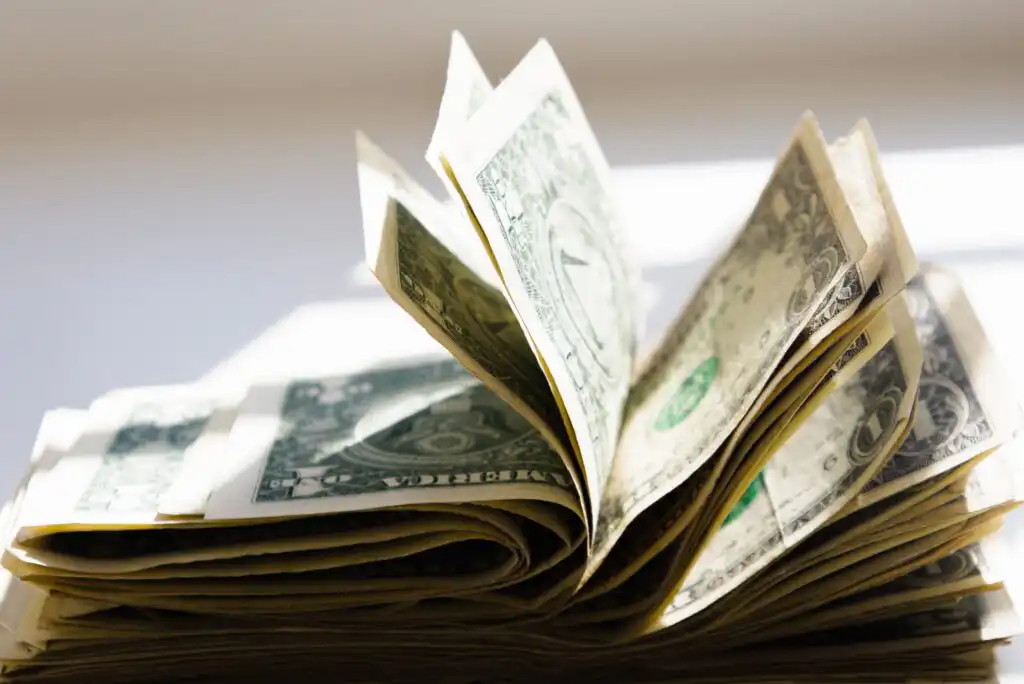 A stack of U.S. one-dollar bills is fanned out, with several bills folded and lifted, set against a bright, sunlit background.