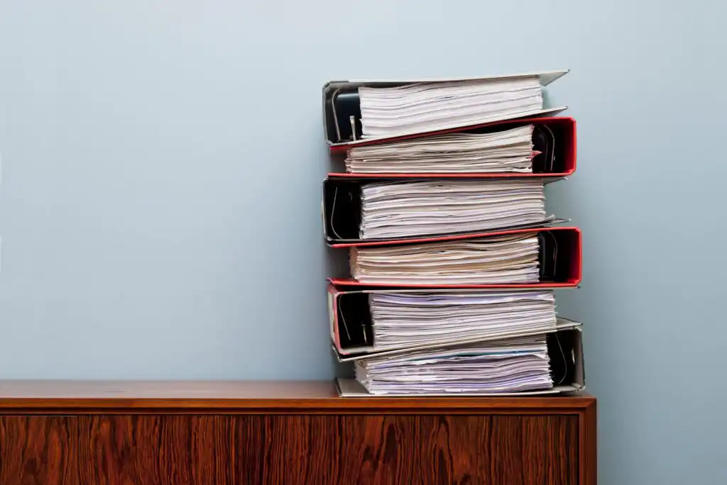 A tall stack of overstuffed binders filled with loose papers sits on top of a wooden cabinet against a plain light blue wall.