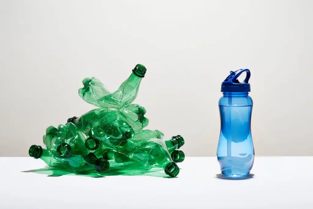 A pile of crushed green plastic bottles sits on the left, while a single reusable blue water bottle stands upright on the right against a plain white background.