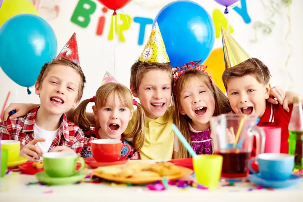 Five smiling children wearing party hats sit together at a decorated table with balloons, colorful cups, plates, and snacks, celebrating a birthday party. Confetti is scattered on the table.