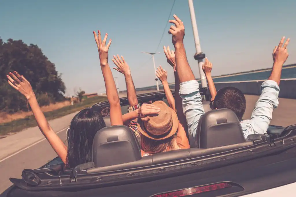 Four people ride in a convertible with the top down, arms raised joyfully in the air, driving along a sunny road near water and trees. The atmosphere appears carefree and adventurous.