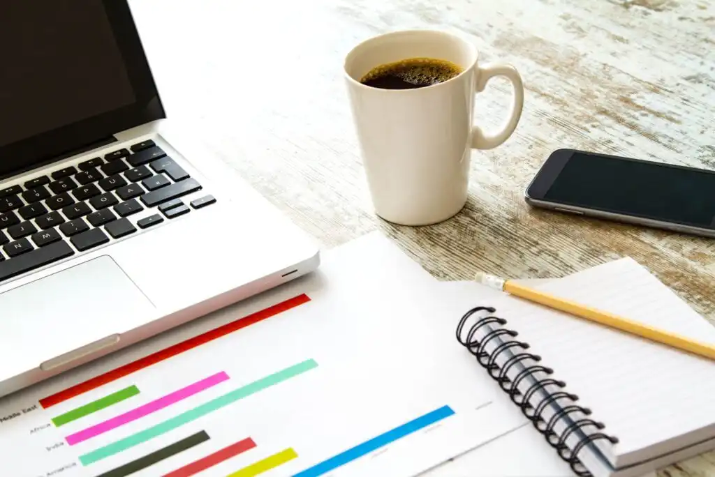 A workspace with a laptop, a cup of coffee, a smartphone, a spiral notebook, a pencil, and documents showing colorful bar graphs on a wooden table.