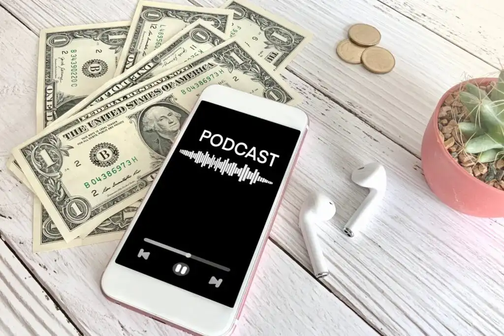 A smartphone displaying a podcast app lies on a white wooden surface with dollar bills, coins, wireless earbuds, and a small potted cactus nearby.
