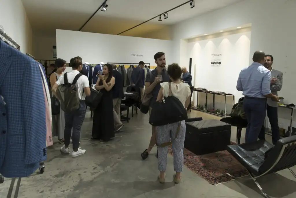 A group of people in business casual attire interact and browse clothing racks in a modern, well-lit showroom with white walls and contemporary decor.