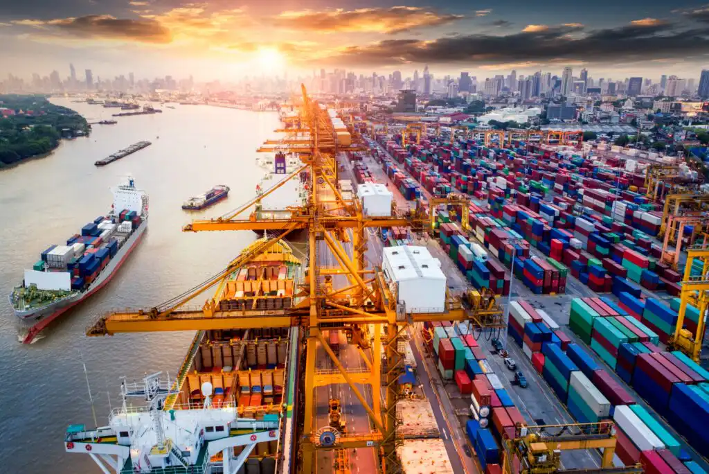 A busy shipping port at sunset with large cranes, stacked cargo containers, and ships docked along the waterfront. A city skyline is visible in the background under a dramatic sky.