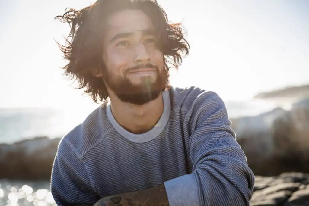 A young man with medium-length wavy hair and a beard smiles while sitting outdoors near the water, wearing a striped long-sleeve shirt. Sunlight shines behind him, creating a bright, relaxed atmosphere.