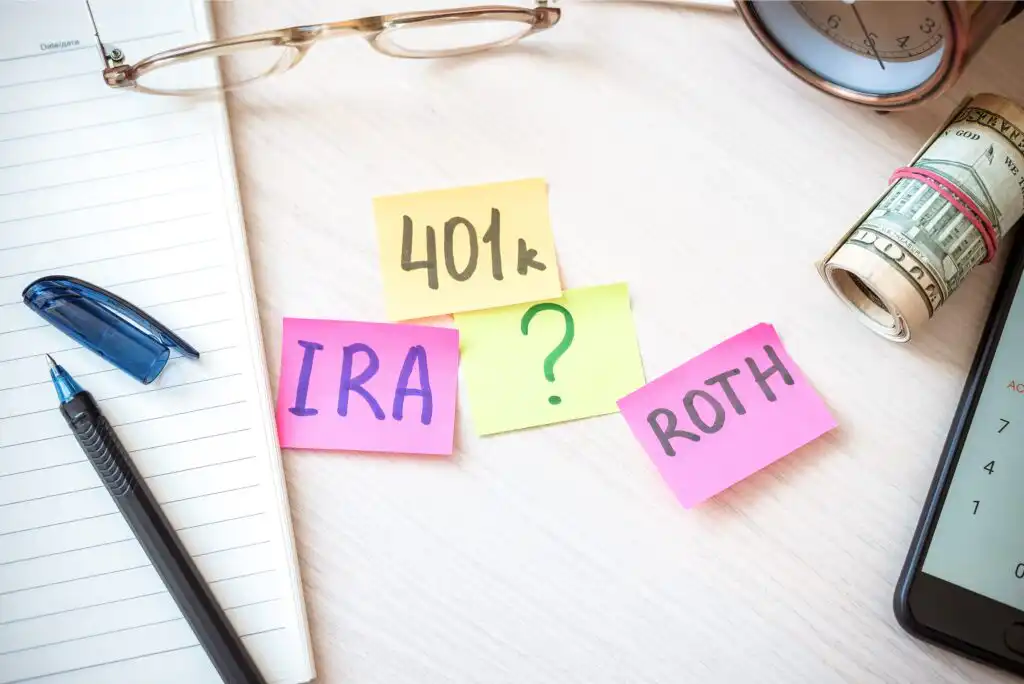 Sticky notes labeled “401k,” “IRA,” “ROTH,” and a green question mark lie on a desk with a pen, notebook, glasses, calculator, and rolled cash, symbolizing retirement savings choices.