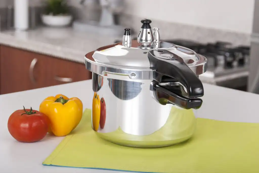 A shiny metal pressure cooker with a black handle sits on a yellow-green mat next to a red tomato and a yellow bell pepper on a white kitchen countertop.