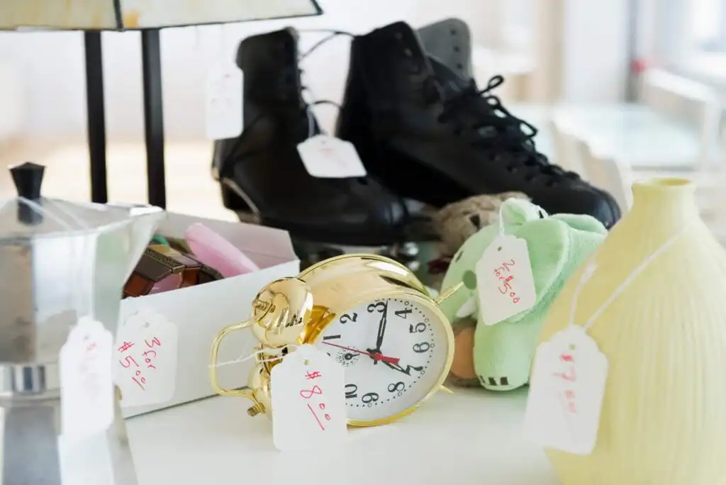 A variety of items with price tags sit on a table, including a gold alarm clock, black boots, a green plush frog, a yellow vase, and a coffee maker, suggesting a yard sale or thrift shop setting.