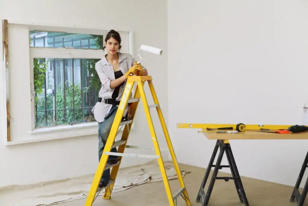 A woman stands on a yellow ladder holding a paint roller in a room being renovated. A large window is behind her, and a workbench with tools is to the right. The floor is covered with protective sheets.