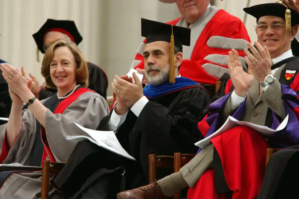 Three people in academic regalia sit and clap at a graduation ceremony. The woman on the left and two men on the right all wear gowns and caps, appearing joyful and engaged in the event.