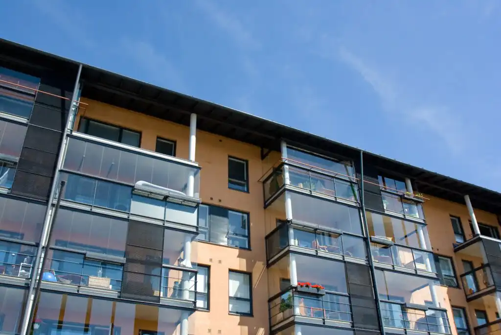 A modern apartment building with multiple glass balconies and a light-colored facade, viewed from below against a clear blue sky. Some balconies have plants and outdoor furniture.