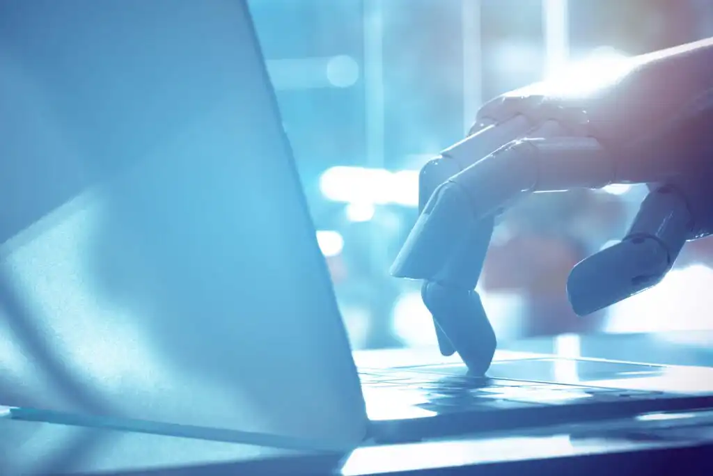 A robotic hand is typing on a laptop keyboard in a brightly lit, modern setting, symbolizing artificial intelligence and technology.