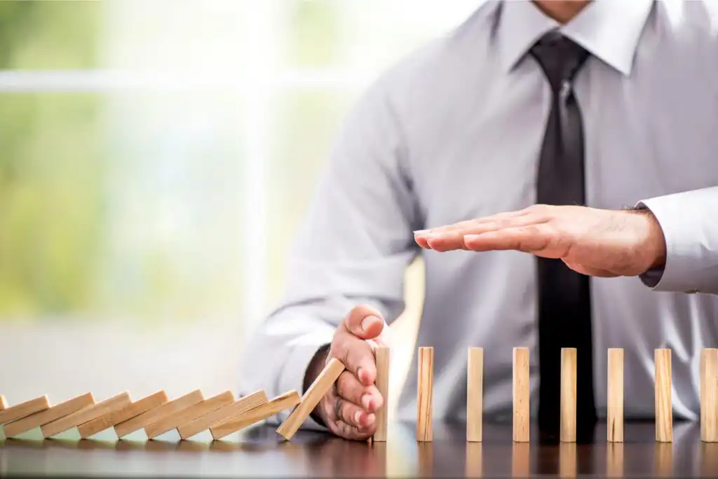 A person in business attire uses their hand to stop falling wooden dominoes on a desk, preventing the remaining upright dominoes from toppling.