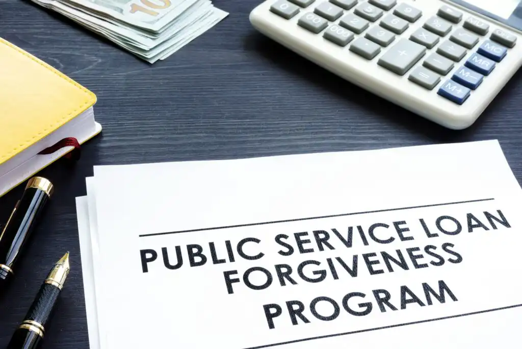 A document titled Public Service Loan Forgiveness Program sits on a desk beside a calculator, a stack of cash, a yellow notebook, and two pens.