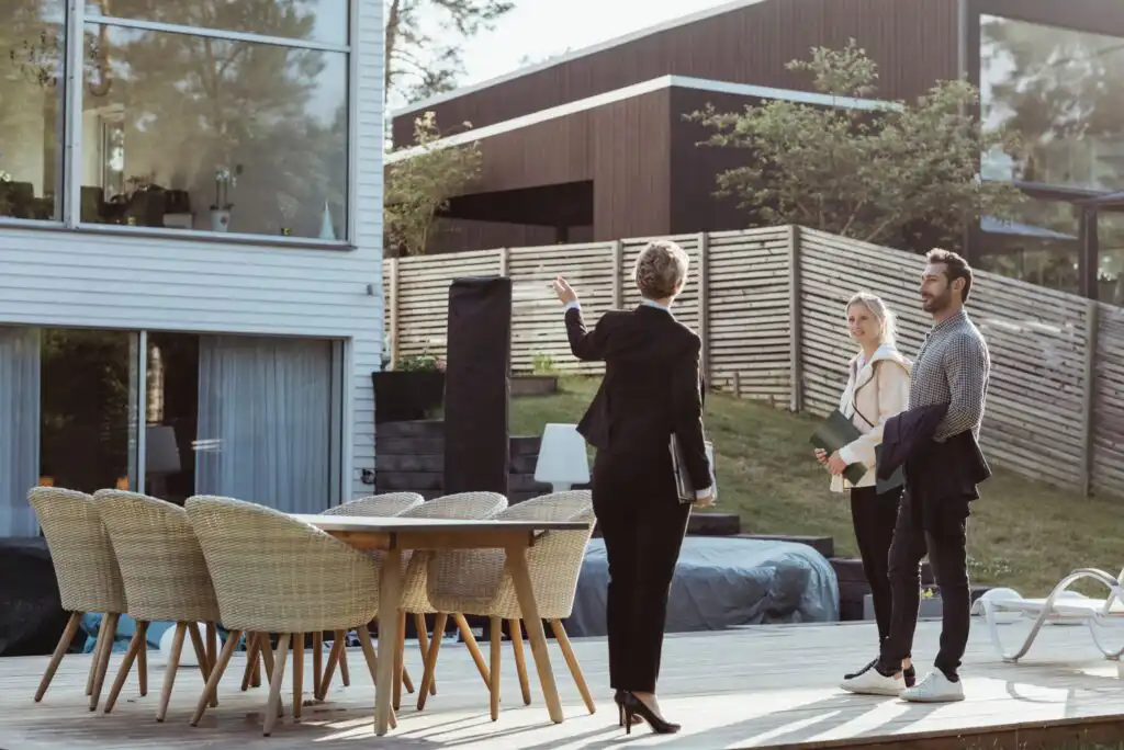 A real estate agent shows a modern house with a wooden deck and outdoor dining set to a man and woman standing outside, holding documents and listening attentively.