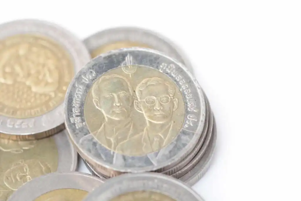 A close-up of stacked Thai ten-baht coins, showing two male figures on the front of the top coin, with Thai script around the edge, on a white background.