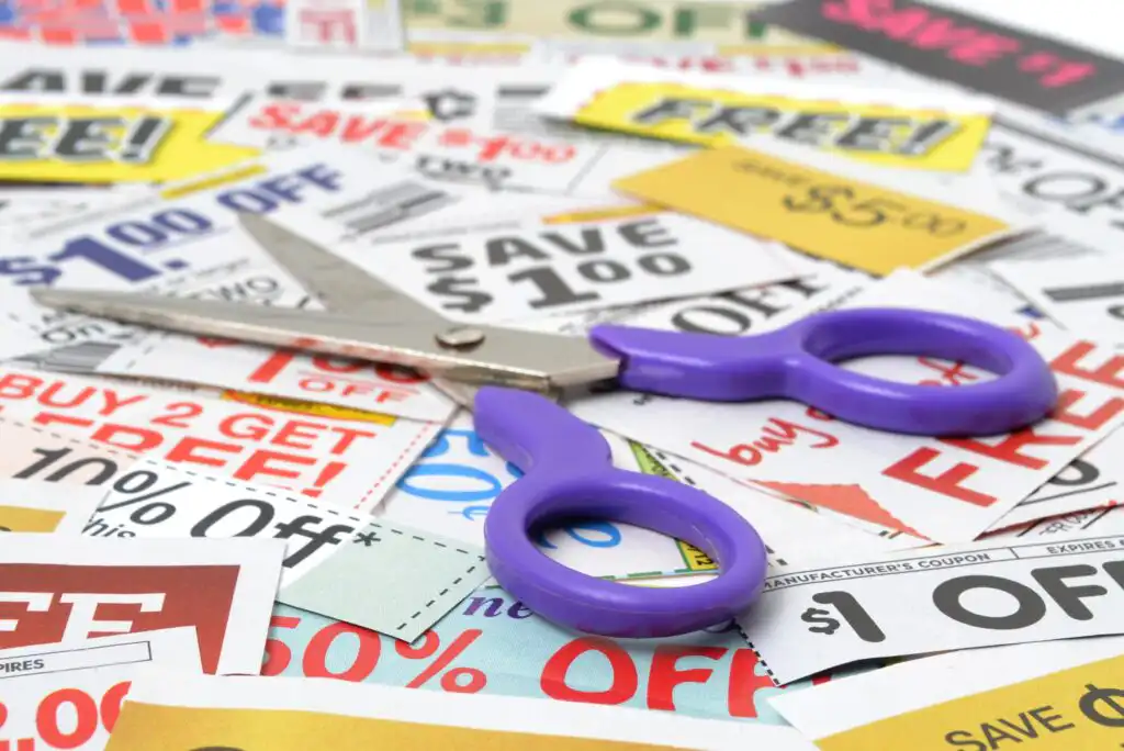 A pair of purple-handled scissors lies on a scattered pile of colorful coupons offering various discounts, such as “$1 off,” “save $1.00,” and “buy 2 get 1 free.”.