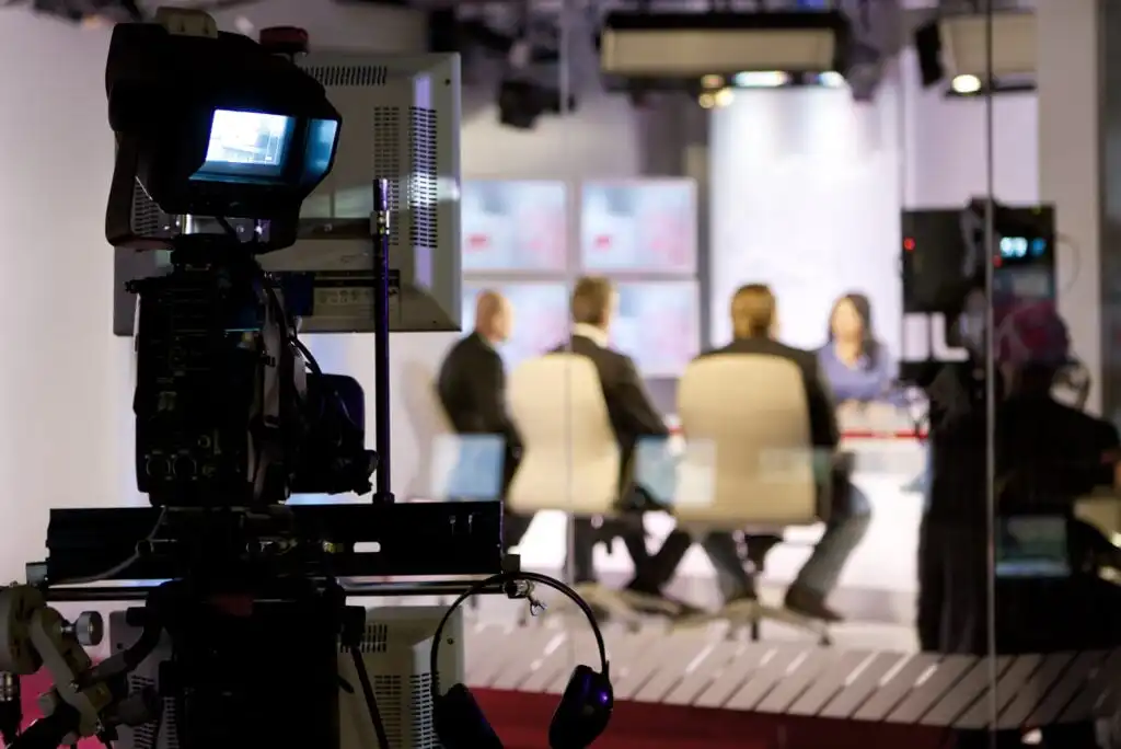 A television studio scene with a video camera in the foreground, focused on four people sitting around a table, engaged in discussion under bright studio lights.