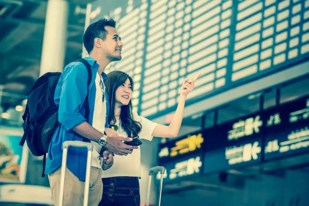 A smiling young couple with luggage looks up at a large digital departures board in an airport; the woman is pointing at the board while the man stands beside her holding a smartphone.