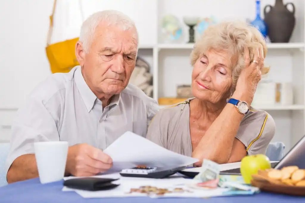 An elderly couple sits at a table looking worried while reviewing papers and bills. A calculator, money, coins, and a cup are on the table, suggesting they are concerned about finances.