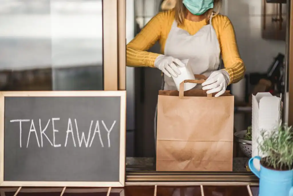A person wearing a mask, gloves, and apron places a takeout container into a brown paper bag at a takeaway window. A chalkboard with TAKEAWAY written on it is visible in the foreground.